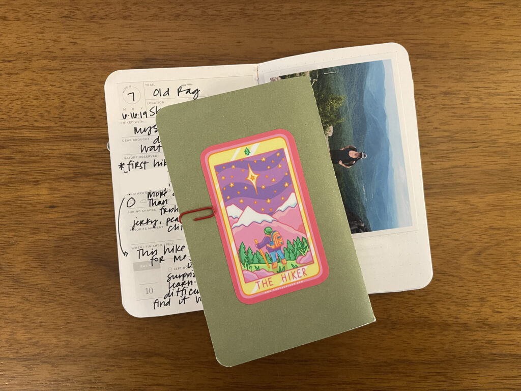 The back of one of my Hike Passports with a mostly pink, purple, and yellow sticker of a hiker setting off through trees into snow-capped mountains.