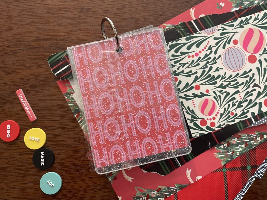 Front cover of my holiday Build-A-Bear album on top of a stack of different holiday-themed gift wrap scraps. The cover is clear with glitter. The title page says "HO HO HO" many times in red and pink. 