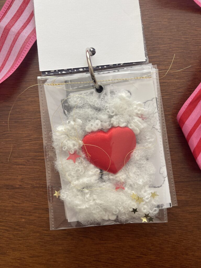 A clear pocket filled with stuffing and a heart from Build-A-Bear, pieces of gold thread, and red and gold star-shaped glitter, stitched together across the top with gold thread. 