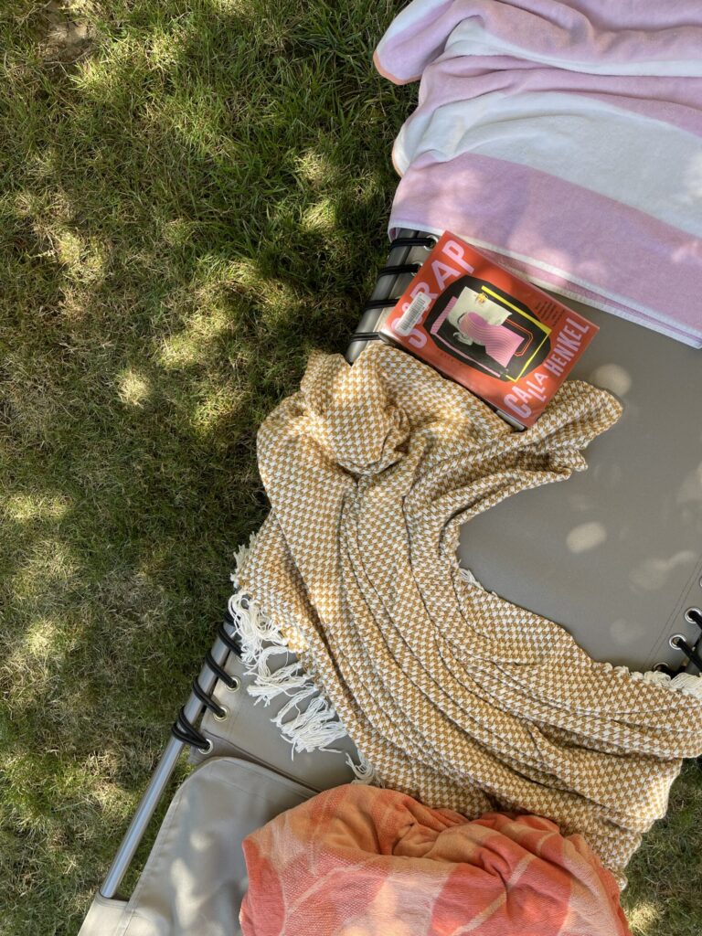 A library book, blanket, and beach towels sitting on my outdoor lounge chair, beneath a tree out front. 
