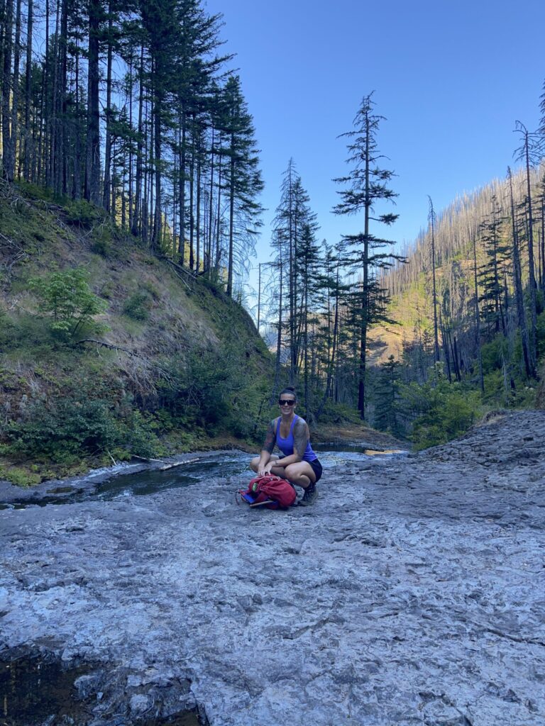 Me, smiling at the camera while crouched in a squat behind my pack on the large, flat rocks at Twister Falls.