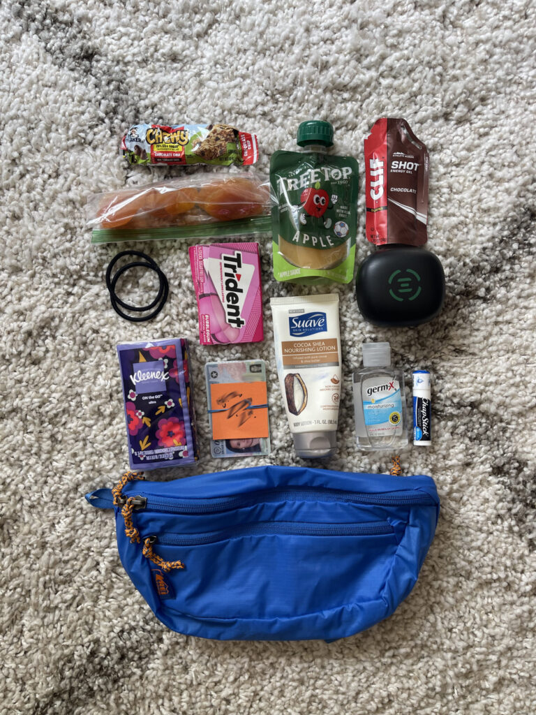 My bright blue REI fanny pack, with bright orange accents, laid out on a rug alongside the items I keep inside it for easy and quick access while hiking—small snacks, hair ties, gum, Kleenex, hand lotion, hand sanitizer, chapstick, earbuds charging case, ID. 