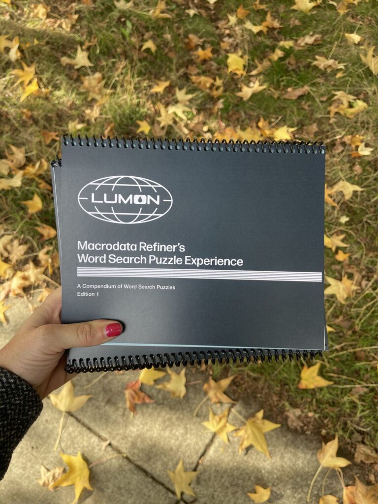 My disembodied hand holding five Lumon/Severance-themed and -branded spiral-bound word search puzzle booklets. The Lumon logo and booklet title text are in white against a navy blue background. Modeled after documents used on the show, the cover reads, "Macrodata Refiner's Word Search Puzzle Experience. A Compendium of Word Search Puzzles. Edition 1."