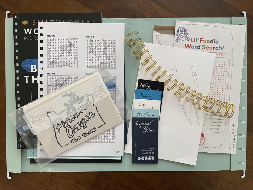 An open file folder, lying flat on a wooden table, holding a variety of examples of different paper colors, weights, and finishes types; and binding that I like. 