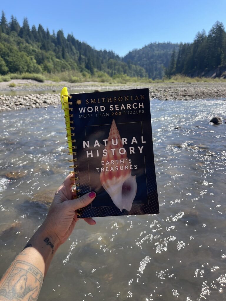 My disembodied hand holding up a Smithsonian "natural history" themed word search puzzle book in front of a river. 