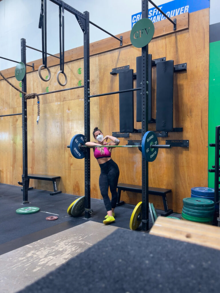 Me, masked, leaning on a racked barbell at the gym, smiling under my mask at the camera. 