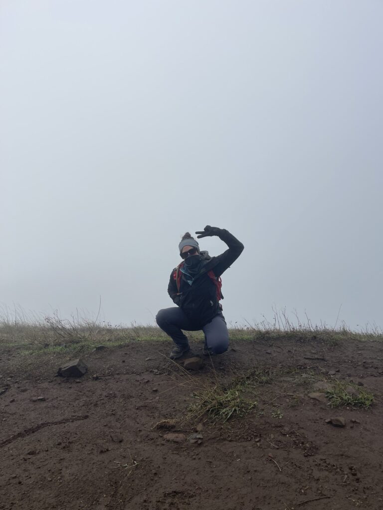 Me, crouched down in front of the fog, giving the camera the peace sign with one gloved hand, at the summit of Dog Mountain.