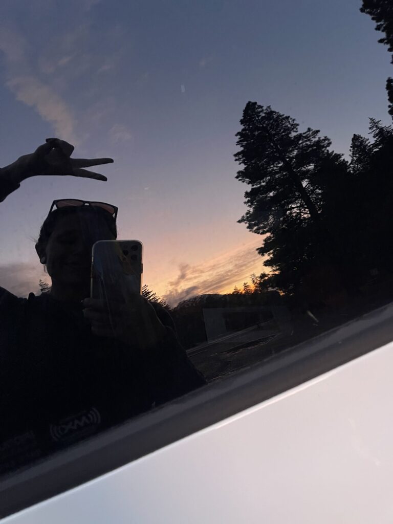A selfie I took of my reflection in my car window, with the orange pre-sunrise in the background. 