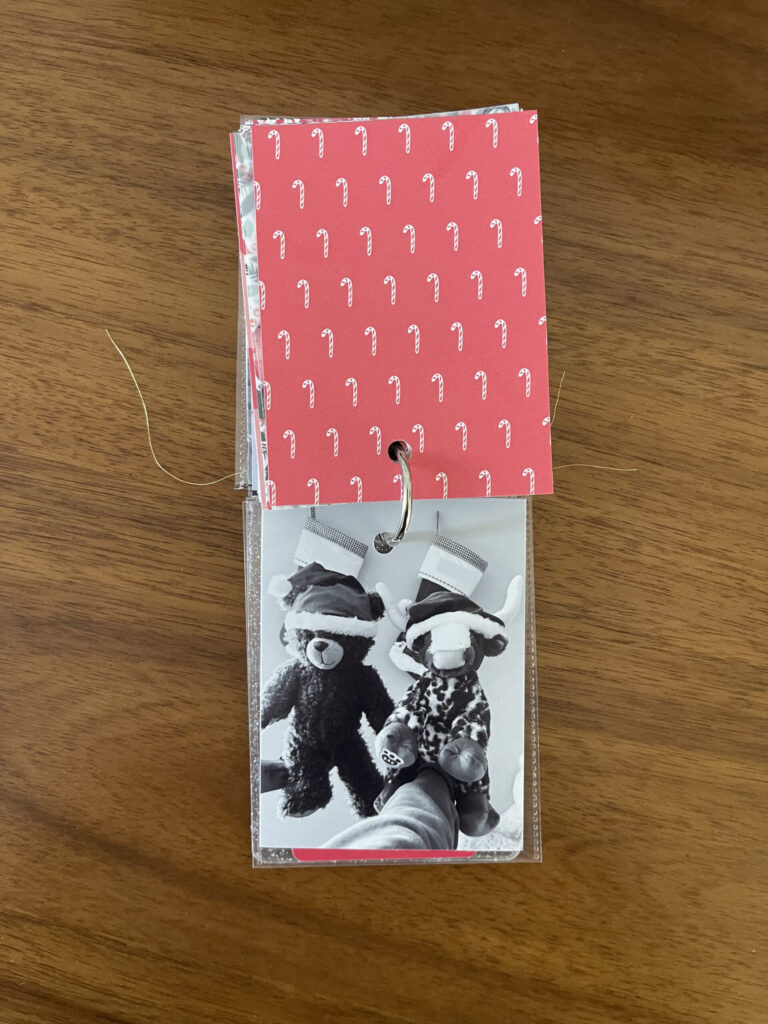2025: red paper with a repeating candy cane pattern on the top page, a black-and-white photo of two outstretched hands, each holding a Build-A-Bear stuffed animal wearing a Santa hat, in front stockings hanging in the background.