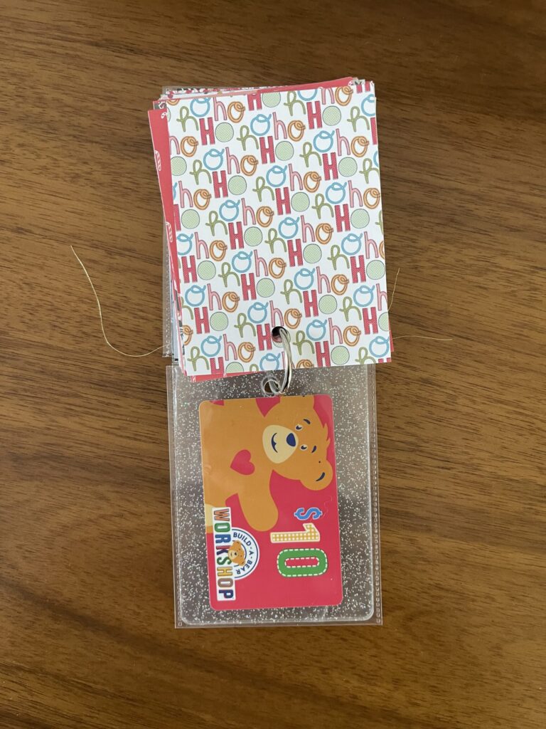 A patterned piece of paper with "ho ho ho" on it on top and a Build-A-Bear gift card in a plastic sleeve on the bottom. 