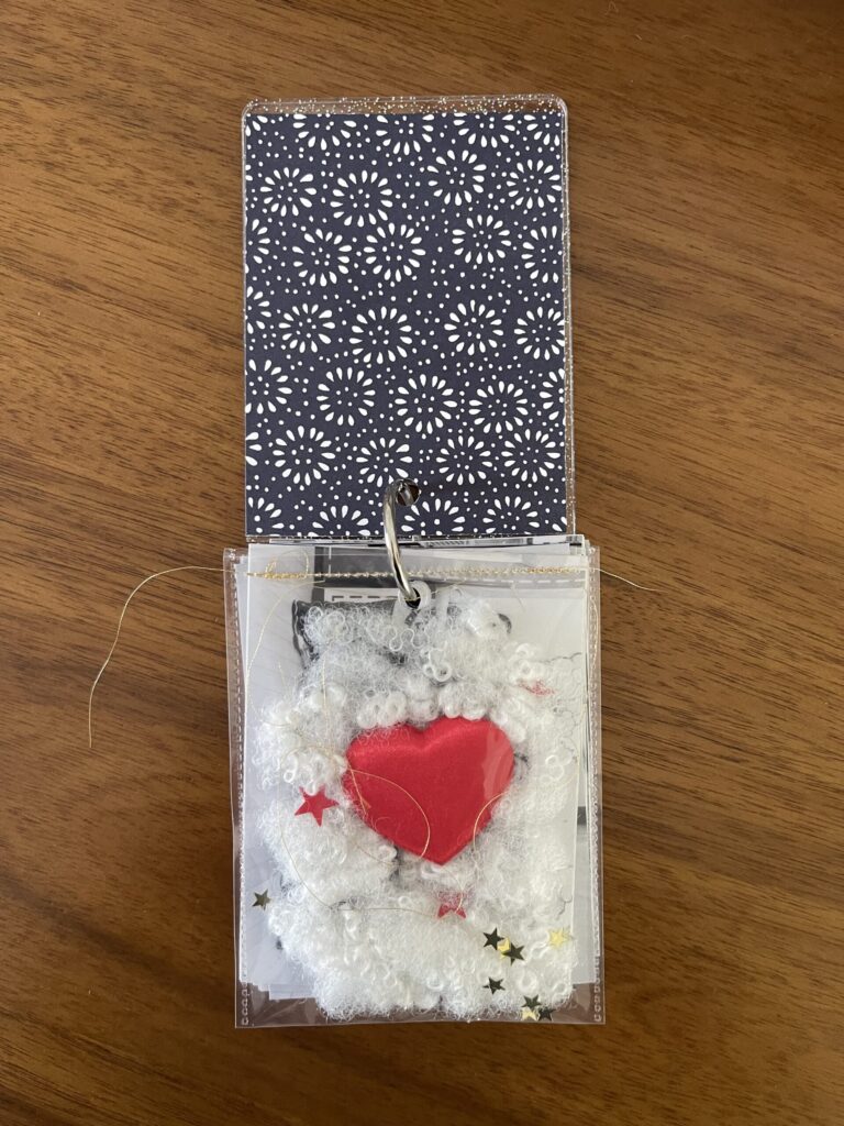 A clear pocket filled with stuffing and a heart from Build-A-Bear, pieces of gold thread, and red and gold star-shaped glitter, stitched together across the top with gold thread. 