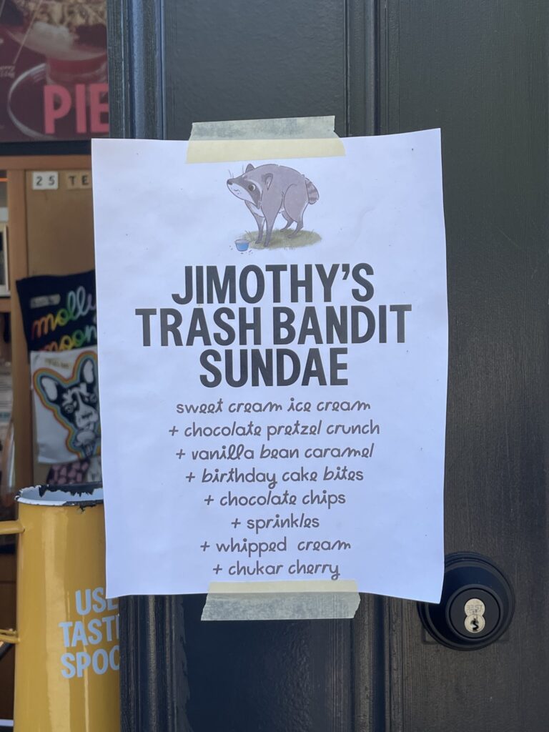 Paper taped to Molly Moon's ice cream stand on the Seattle waterfront detailing the ingredients in Jimothy's Trash Bandit Sundae: sweet cream ice cream, chocolate pretzel crunch, vanilla bean caramel, birthday cake bites, chocolate chips, sprinkles, whipped cream, chukar cherry.