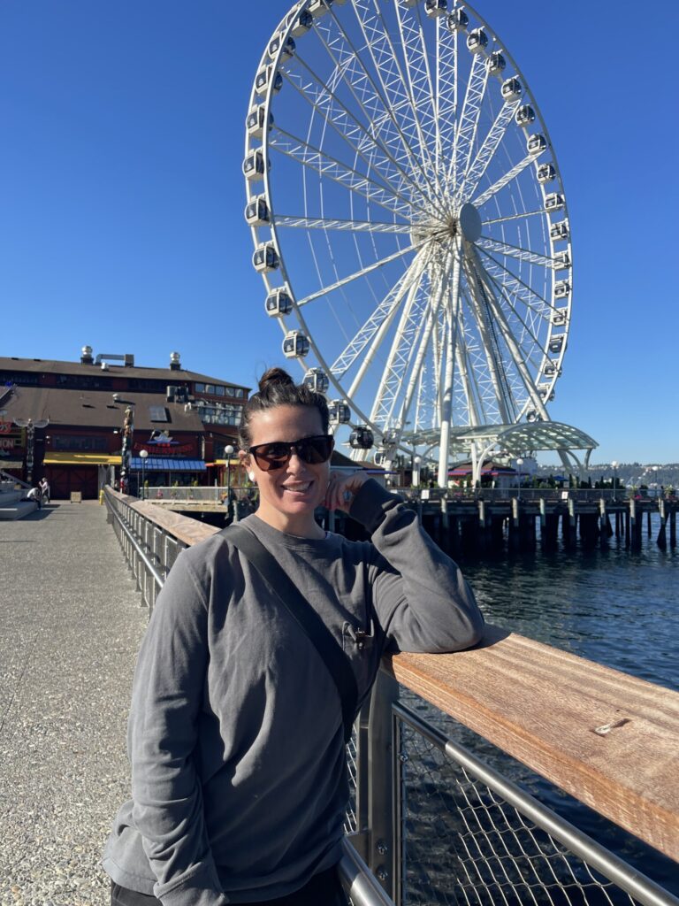 Me, leaning against a chest-high fence along the waterfront, with Elliot Bay to my left and a Ferris wheel directly behind me. 