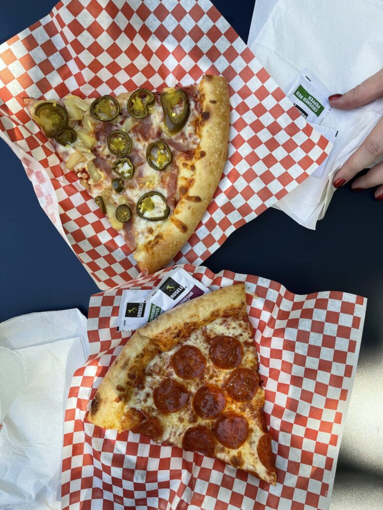 Overhead view of two large slices of pizza on red and white checkered paper atop paper plates on a navy blue table. My slice has cheese and pepperoni, my friend's has cheese, peppers, ham, and pineapple. 
