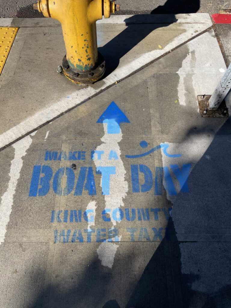 "Make it a boat day, King County water taxi" stenciled in bright blue on a wet sidewalk near the Seattle waterfront.
