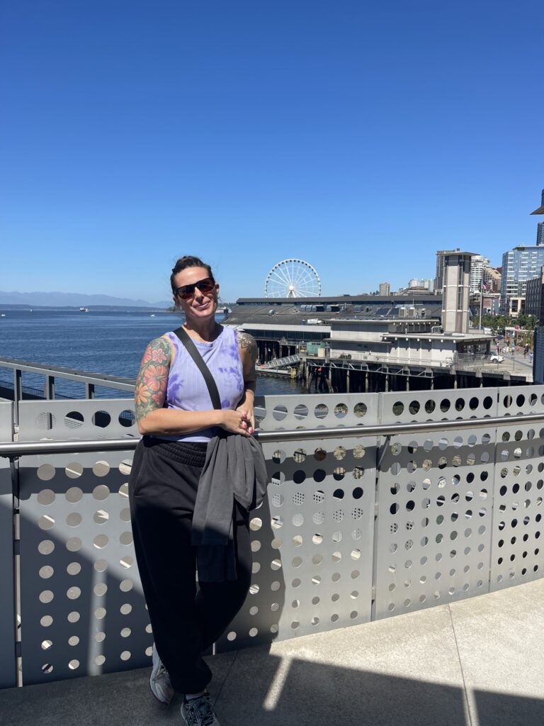Me, standing against a railing while waiting to board the ferry from Seattle to Bainbridge Island. Elliott Bay and sky are bright blue, and a Ferris wheel is in the background. 