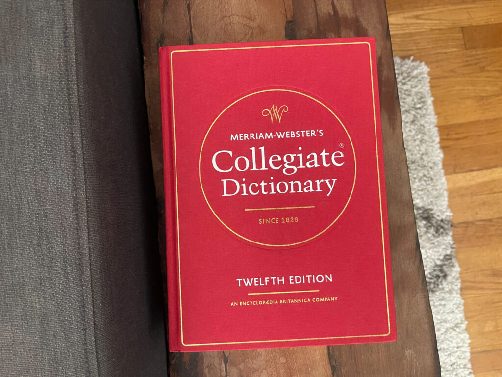Merriam-Webster's Collegiate Dictionary, 12th Edition face up on a wooden side table. The cover is red cloth outlined in gold. 
