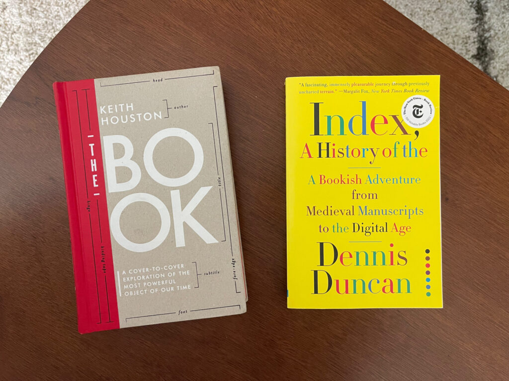 The books "The Book" and "Index, A History of the" face up on a wooden table.