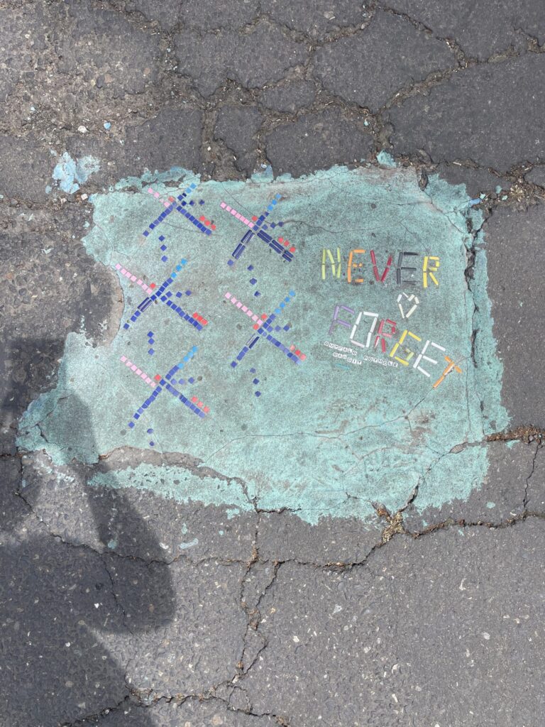 A pothole in a SE Portland street filled by a street artist to look like the iconic old PDX carpet. Tiles spell out "NEVER FORGET" alongside a heart. Friendship bracelet beads spell out "BUFFALO POTHOLE BANDIT"