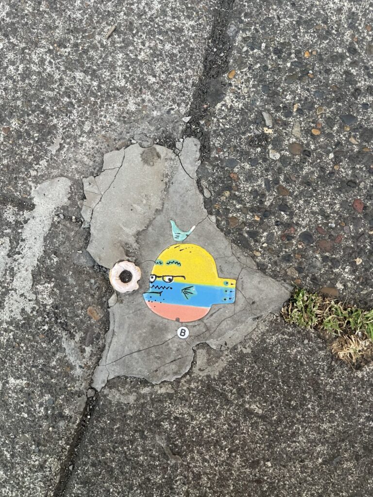 A tile of a colorful fish with a small bird atop its head and a friendship bracelet bead of the letter B. 